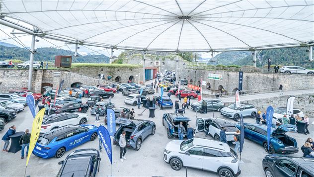 Eine Autoausstellung unter einem großen Zelt mit zahlreichen Fahrzeugen und Besuchern. Im Hintergrund sind historische Mauern und eine grüne Landschaft zu sehen.